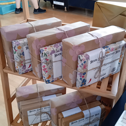 Several wrapped gift boxes tied with twine and labeled 'Book-Love Mystery Box' displayed on a wooden shelf in a store or event setting.