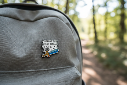 A Black and white pin with a sword design that says Current side quest brought to you by ADHD on a backpack