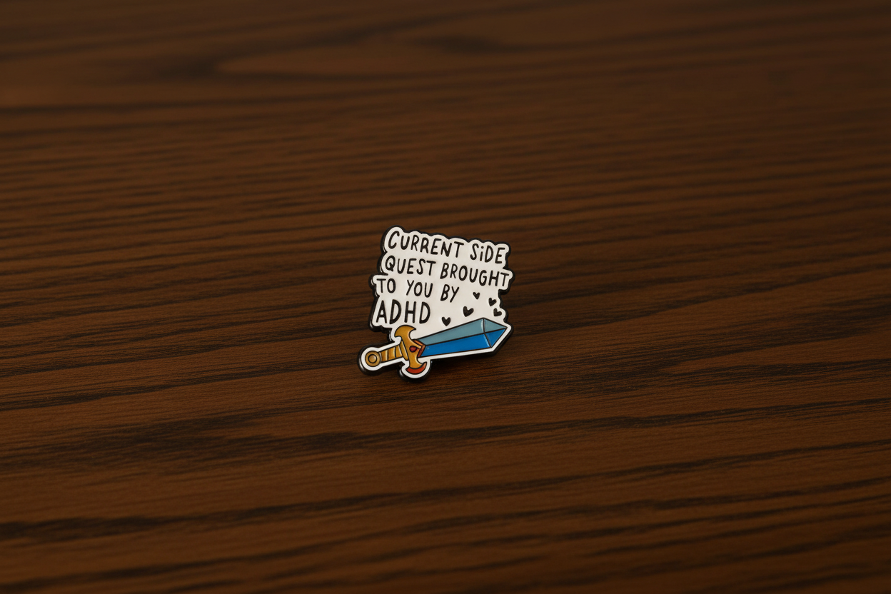 A Black and white pin with a sword design that says Current side quest brought to you by ADHD on a dark oak table