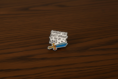 A Black and white pin with a sword design that says Current side quest brought to you by ADHD on a dark oak table
