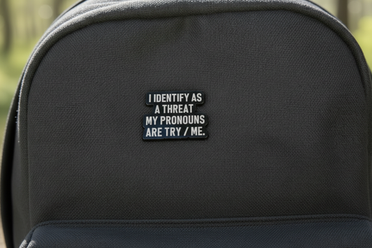 A Black and White Enamel Pin Badge that Says I identify as a threat, my pronouns are try/me. This is on a backpack