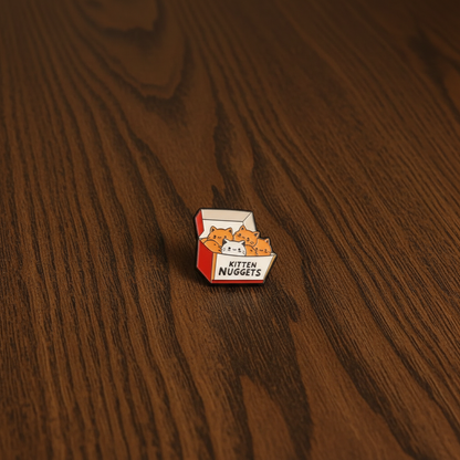 A cute pin badge of Kittens in a chicken nugget box that says Kitten Nuggets. The pin is on a dark oak table