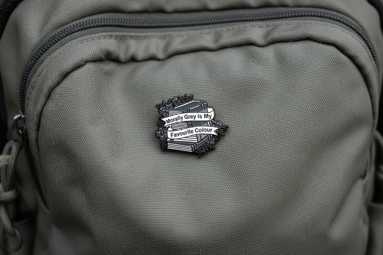 An enamel pin badge of a greyscale stack of books that says "Morally Grey is my favourite Colour"