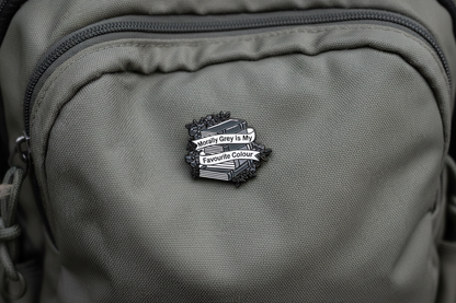 An enamel pin badge of a greyscale stack of books that says "Morally Grey is my favourite Colour"
