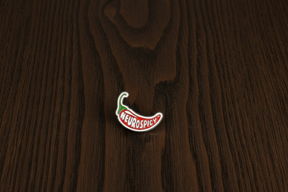 Enamel pin of a chili pepper with 'NEUROSPICY' text on a dark oak table