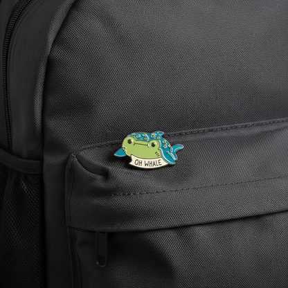 Small whale-shaped enamel pin with 'OH WHALE' text on a backpack