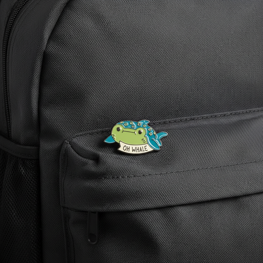 Small whale-shaped enamel pin with 'OH WHALE' text on a backpack